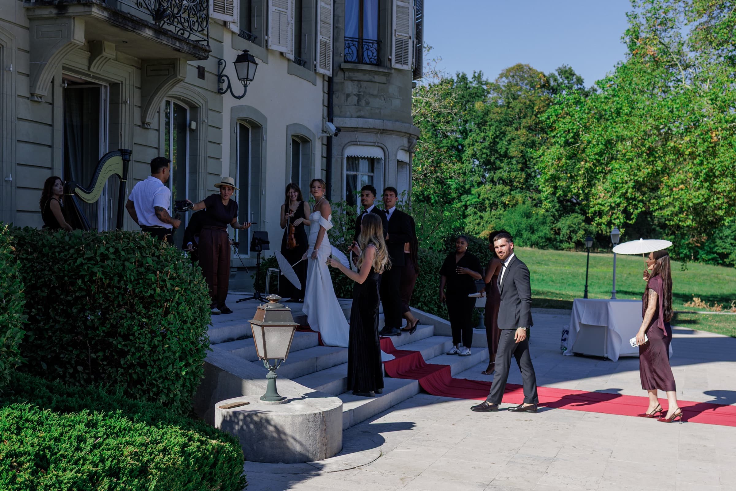 Arrivée sur les marches du château : tapis rouge, harpiste, violoniste, mariée à l'éventail et invités en tenue de soirée sous des ombrelles blanches — Château de Crassy