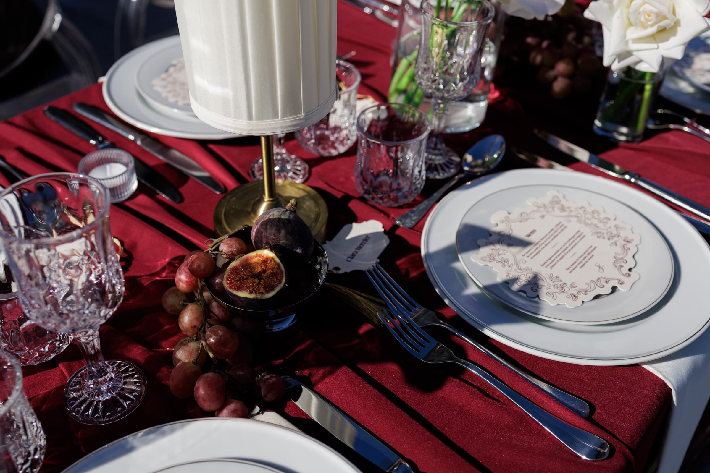 Détail d'art de la table : velours bordeaux, cristal taillé, figues ouvertes et grappes de raisin, menu enluminé et assiette à liseré d'argent — Château de Crassy