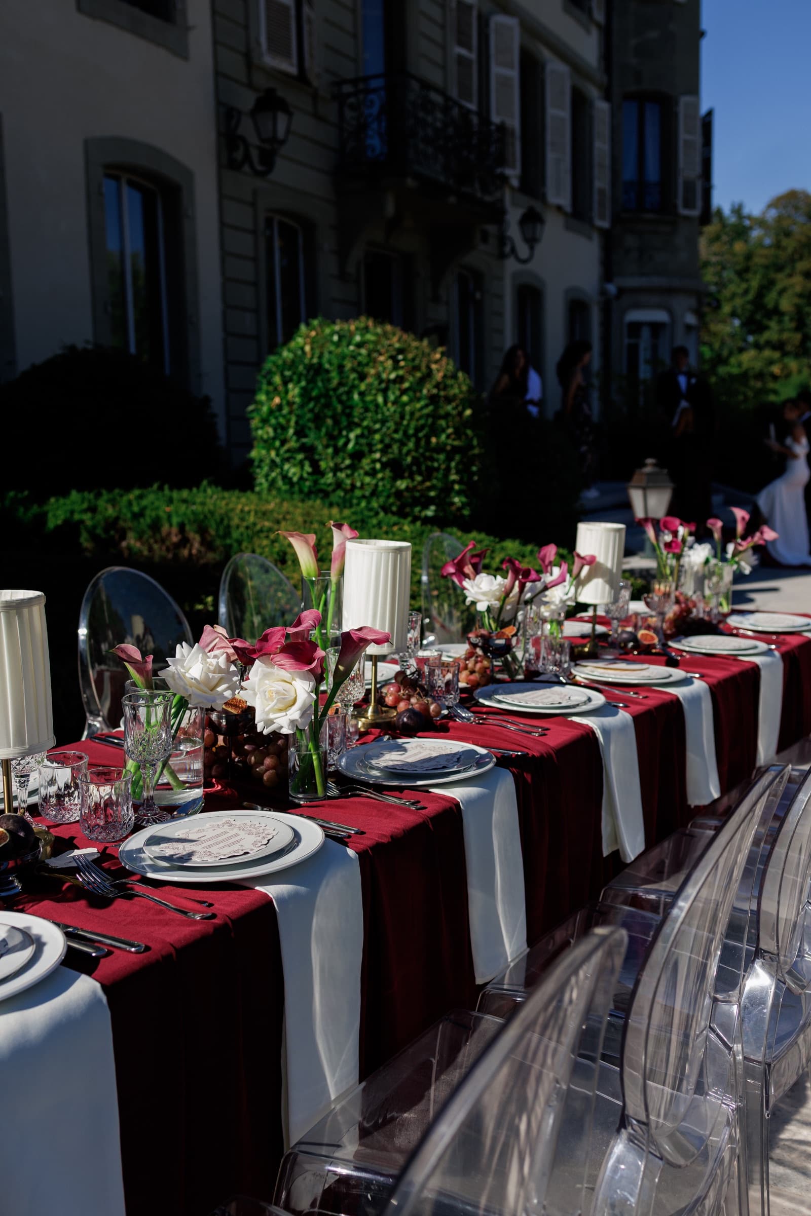 Longue table de banquet dans le parc : chemins de velours bordeaux, chaises fantômes de cristal, arums grenat et roses blanches, lampes à abat-jour — Château de Crassy