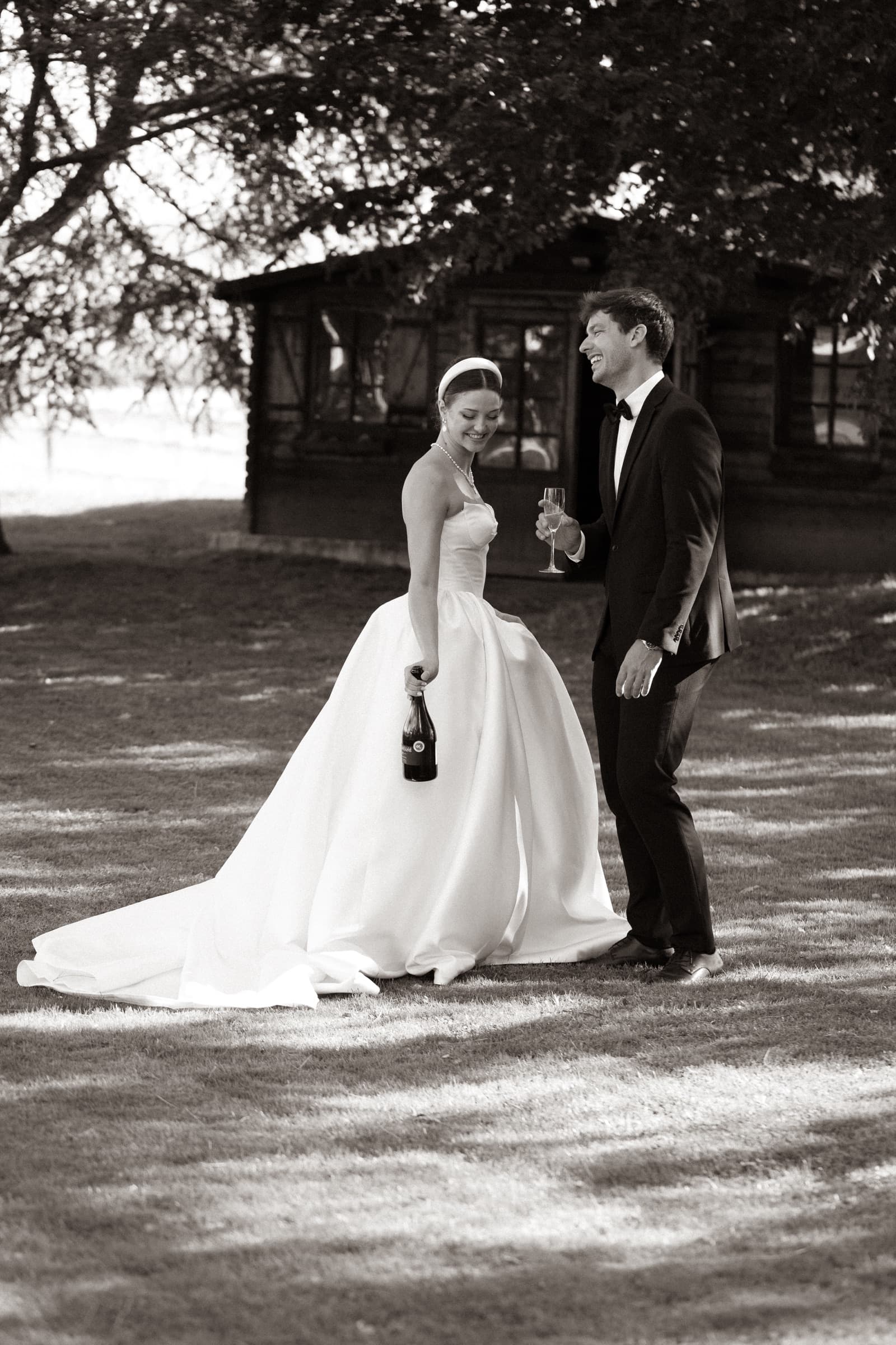 Robe Atelier Pointillé en pied, mariée et marié riant devant le chalet en bois, noir et blanc — éditorial mariage Domaine La Capitaine