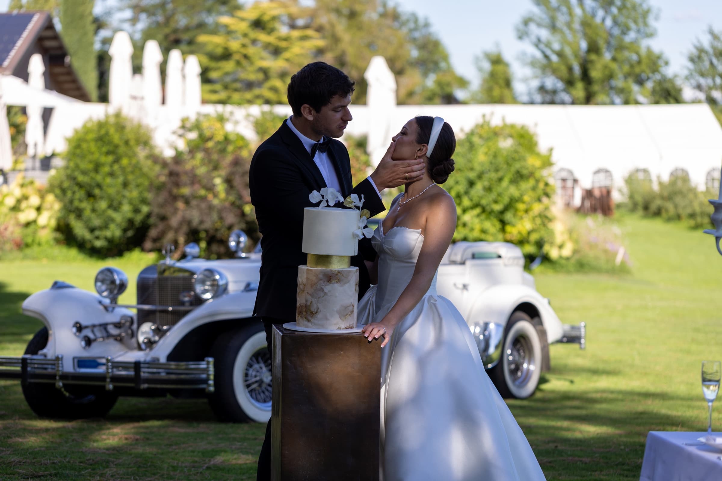 Le couple devant le wedding cake à trois étages d'Universsa Cakes, feuille d'or et orchidées blanches, roadster de collection en arrière-plan — Domaine La Capitaine